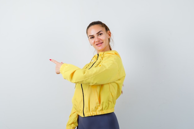 Señorita de chaqueta amarilla apuntando a un lado y mirando alegre, vista frontal.