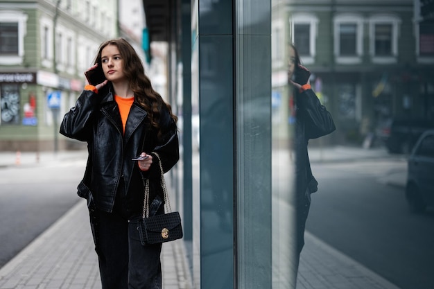 Señorita en chaqueta de cuero caminando en ciudad urbana hablando por teléfono