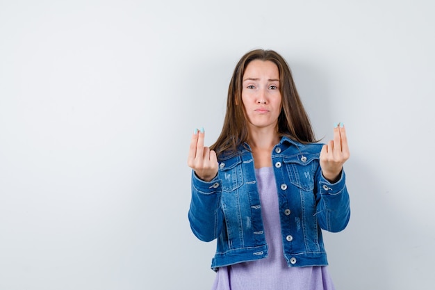 Foto señorita haciendo gesto de dinero en camiseta, chaqueta y con cara de tristeza. vista frontal.