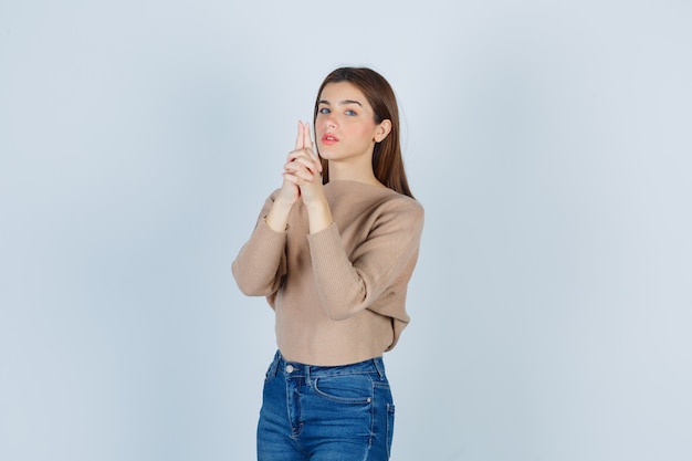 Señorita mostrando gesto de pistola en suéter beige, jeans y mirando confiado, vista frontal.