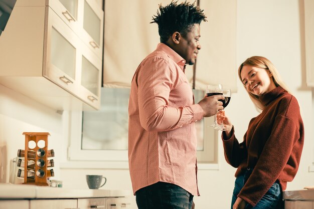 Señorita positiva inclinando la cabeza hacia la derecha y sonriendo mientras bebe vino en la cocina con el joven