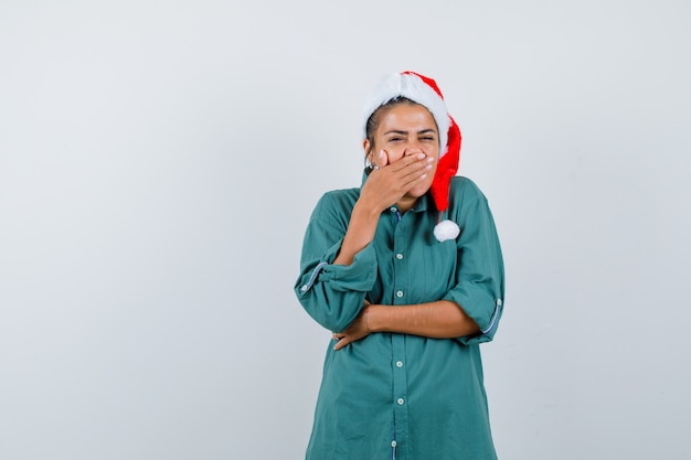 Señorita con sombrero de navidad, camisa con la mano en la boca y mirando ashmed, vista frontal.