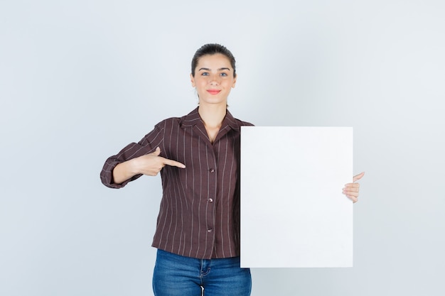 Señorita sosteniendo un cartel de papel, apuntando hacia el lado en camisa, jeans y mirando complacido, vista frontal.