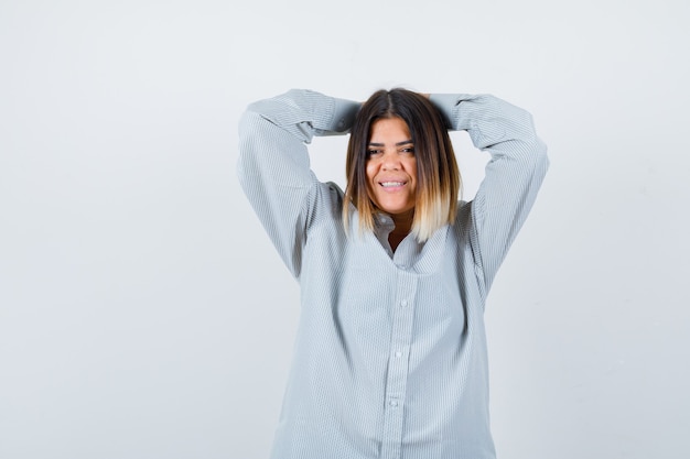 Señorita sosteniendo los dedos entrelazados detrás de la cabeza en camisa de gran tamaño y mirando complacido, vista frontal.