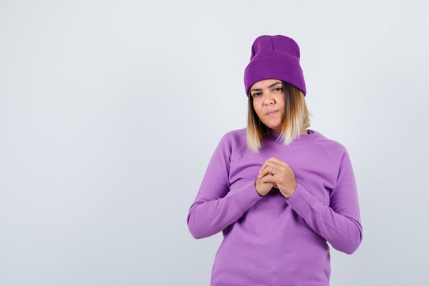 Señorita sosteniendo las manos juntas en el pecho en suéter morado, gorro y mirando enfocado, vista frontal.