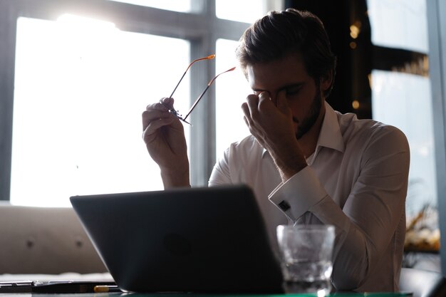 Foto sensación de malestar y cansancio. hombre joven frustrado masajeando su nariz y manteniendo los ojos cerrados mientras está sentado en la computadora portátil en la cafetería.