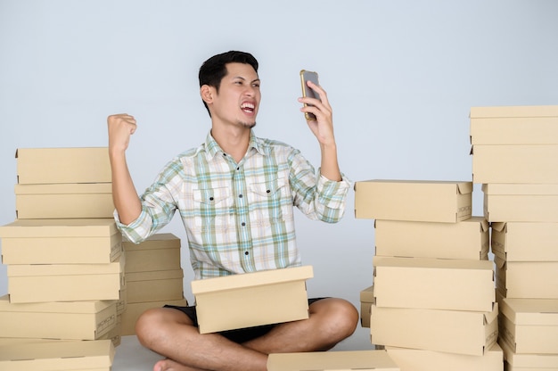 Foto sensación muy satisfecha y cara feliz de hombre asiático mira smartphone con levantar un puño entre muchas cajas con paquetes en una pared blanca.