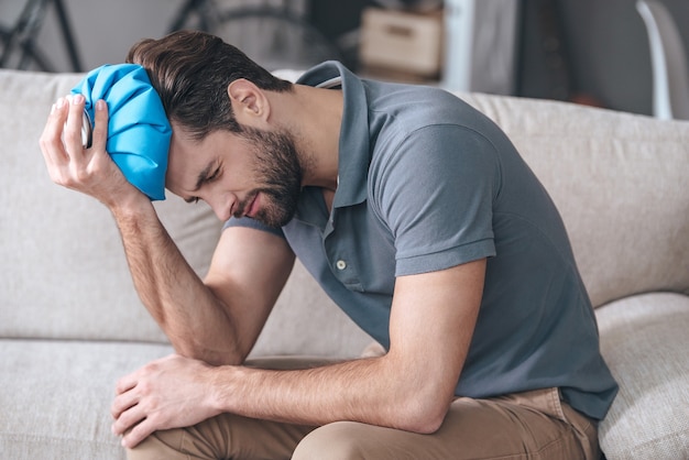 Sensación de un terrible dolor de cabeza. Hombre joven frustrado que sostiene una bolsa de hielo en la cabeza mientras está sentado en el sofá en casa