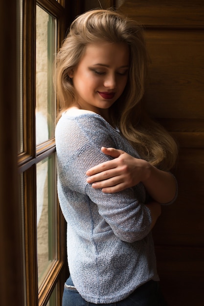 Sensual modelo rubia con maquillaje profesional y cabello largo posando en la ventana