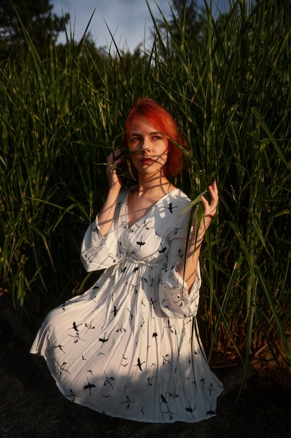 Sensual mujer bonita con el pelo corto y rojo se sienta en el suelo entre la hierba, posa y mira la puesta del sol