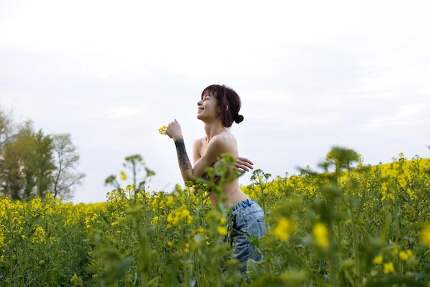 Sensual mujer joven con tatuajes posando en un campo de colza entre flores amarillas en un día soleado.