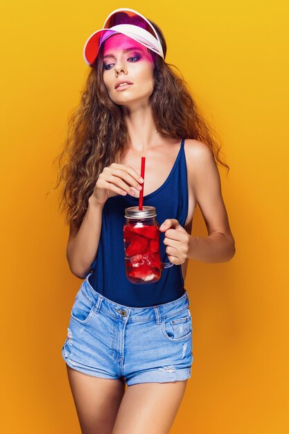 Sensual mujer joven en traje de verano, tapa con jarra con bebida fresca y mientras está de pie en amarillo brillante