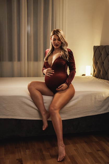 Foto sensual mulher grávida vestindo cueca bodysuit vermelho rubi enquanto relaxa no quarto.