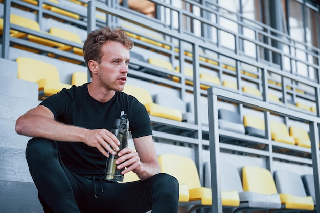 Senta-se com garrafa de água Jovem esportivo de camisa preta e calças ao ar livre durante o dia