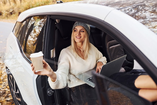 Foto senta-se com o laptop e um copo de bebida. menina tem viagem de outono de carro. automóvel novo moderno na floresta.