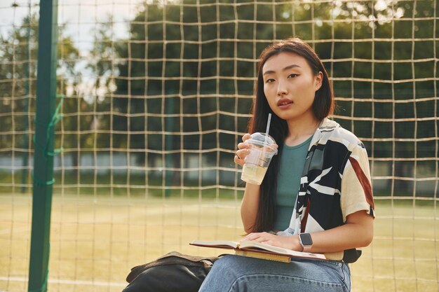 Senta-se contra o campo esportivo a jovem asiática está ao ar livre durante o dia