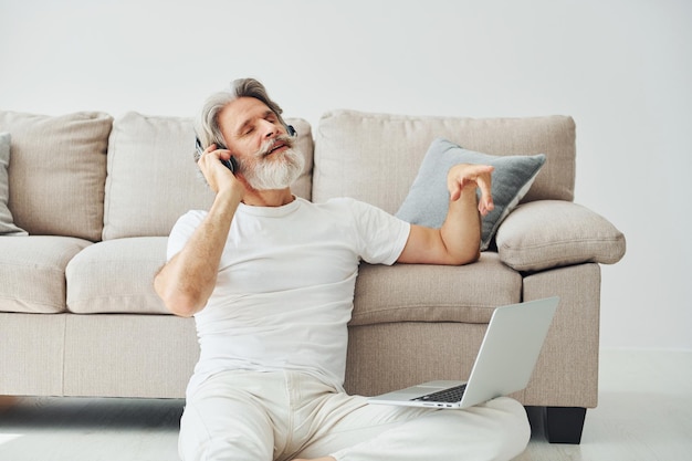 Senta-se no chão perto do sofá com laptop e ouve música Homem moderno elegante sênior com cabelos grisalhos e barba dentro de casa