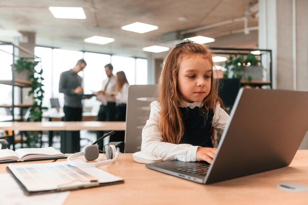 Sentada à mesa com o laptop, a menina está no escritório onde as pessoas trabalham juntas.