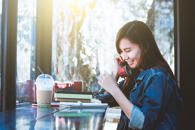 Sentada adolescente de Asia sola que usa el teléfono rojo con la sonrisa en café.