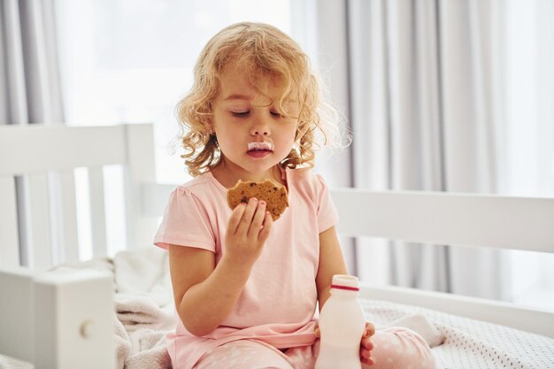 Sentada en la cama comiendo galletas y bebiendo leche Linda niña con ropa informal está en casa durante el día