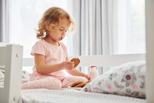 Sentada na cama comendo biscoitos e bebendo leite Menina bonitinha em roupas casuais está dentro de casa durante o dia