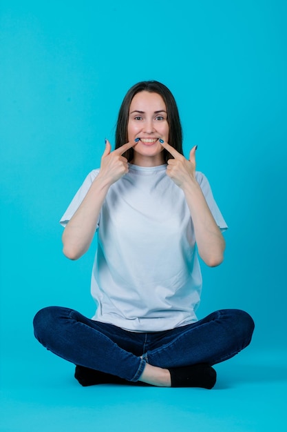 Sentada en el suelo, la chica muestra su sonrisa con los primeros planos sobre fondo azul.