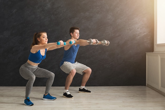 Sentadilla profunda. Pareja de fitness en ropa deportiva haciendo ejercicios de sentadilla, sosteniendo pesas en el gimnasio, espacio de copia