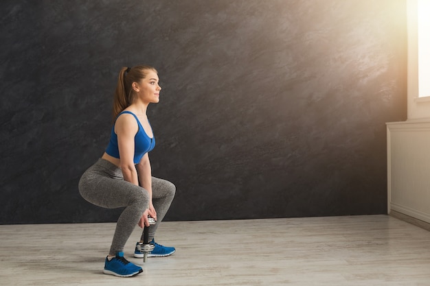 Sentadilla profunda. Vista lateral de la joven y bella mujer en ropa deportiva haciendo ejercicios de sentadillas, sosteniendo pesas en el gimnasio, espacio de copia