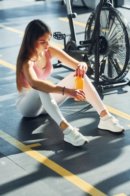 Sentado con una botella de agua Hermosa mujer joven con cuerpo delgado está en el gimnasio