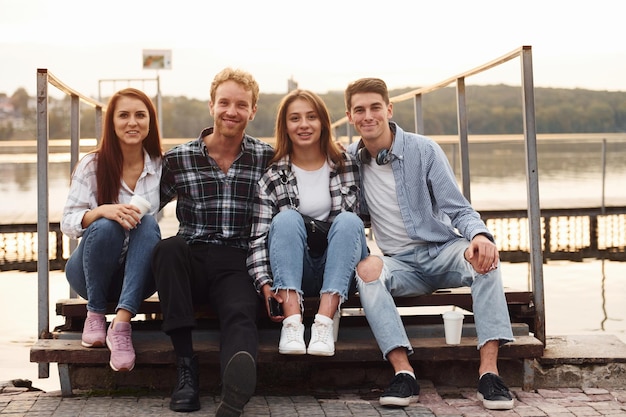 Sentado cerca del lago y abrazándose Grupo de jóvenes amigos alegres que están al aire libre divirtiéndose juntos