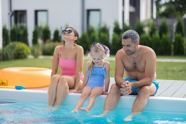 Sentado cerca de la piscina. Los padres y su encantadora hija sentados junto a la piscina en un caluroso día de verano