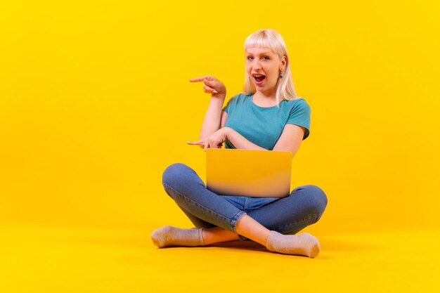 Sentado com um laptop apontando para a garota caucasiana loira esquerda no estúdio em fundo amarelo
