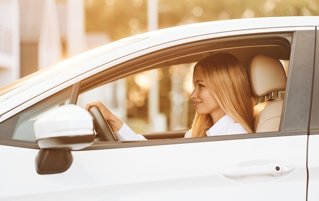 Sentado y conduciendo Mujer joven vestida de blanco está con su coche eléctrico durante el día