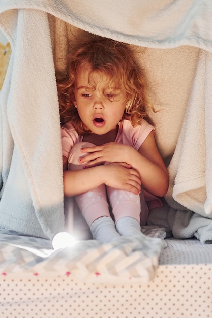 Sentado bajo una manta Linda niña con ropa informal está en el interior de su casa durante el día