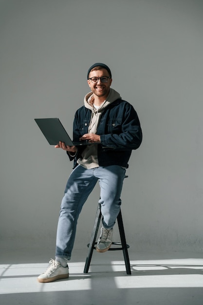 Sentado na cadeira e segurando um laptop, um homem bonito está no estúdio contra um fundo branco.