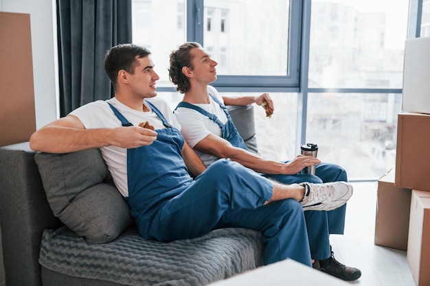 Sentado no sofá Cansado e fazendo uma pausa Dois jovens em uniforme azul trabalhando dentro de casa na sala