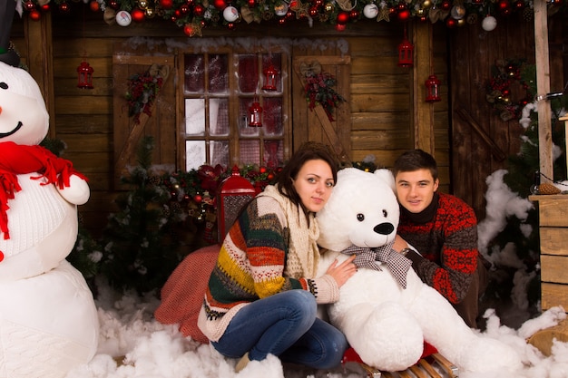 Sentado pareja joven en trajes de invierno abrazando muñeco de oso de invierno blanco en casa de madera decorada con muñeco de nieve grande y varias decoraciones navideñas.