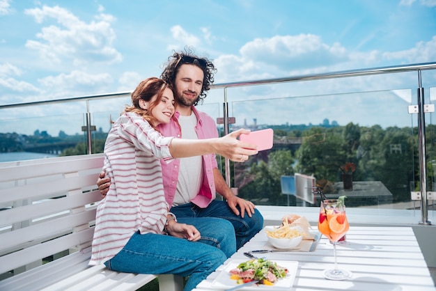 Sentado en la terraza. Mujer sonriente haciendo fotos con su hombre sentado en la terraza durante la cena romántica