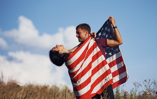 Sente liberdade. Lindo casal com bandeira americana se diverte ao ar livre no campo.