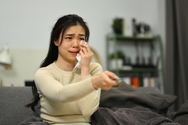 Sentimentale junge Frau weint und wischt sich die Tränen mit Papiertaschentuch ab, während sie sich zu Hause einen dramatischen, traurigen Film ansieht