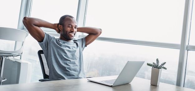 Sentindo alegria. Alegre jovem empresário americano africano está reclinado na cadeira e segurando as mãos atrás da cabeça enquanto olha para a tela com um sorriso largo