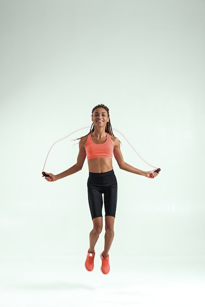 Foto sentindo feliz. comprimento total da bela mulher africana sorridente com corpo perfeito, pular corda durante o exercício no estúdio contra um fundo cinza. equipamento esportivo. estilo de vida ativo