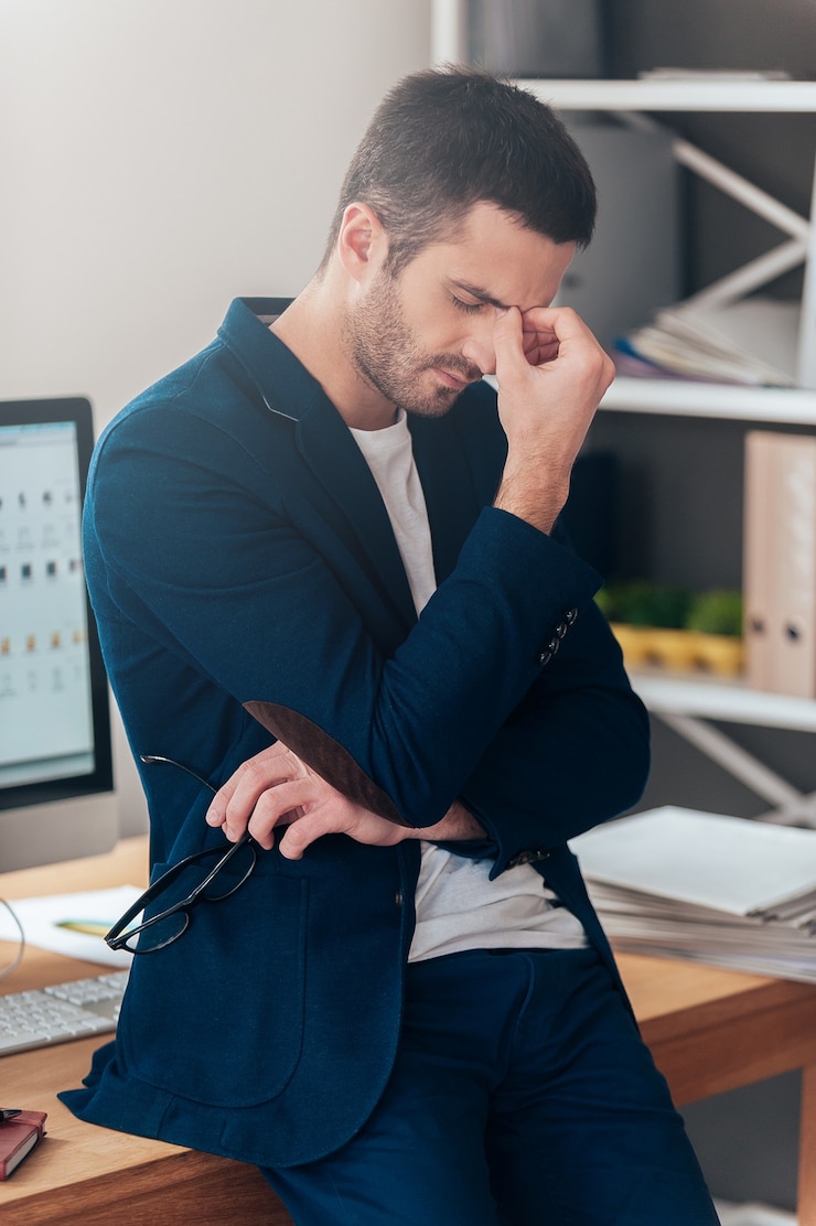 Foto sentindo-se cansado e deprimido. homem jovem frustrado em uma roupa casual elegante, usando óculos e mantendo os olhos fechados, enquanto se inclina na mesa do escritório