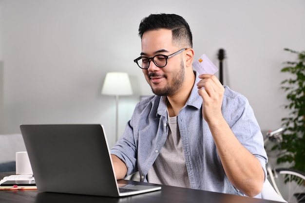 Sentindo-se feliz e confiante. Jovem asiático usando computador portátil segurando cartão de crédito para compras on-line e pagamento em casa.