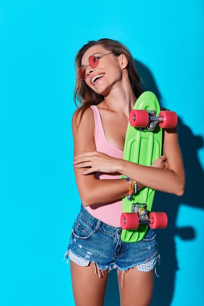 Sentindo-se livre e feliz. Mulher jovem e atraente sorrindo e carregando um skate em pé contra um fundo azul