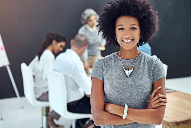 Foto sentindo-se positivo em relação à minha nova equipe retrato de uma empresária em pé com os braços cruzados na sala de reuniões enquanto um colega faz uma apresentação em segundo plano