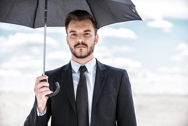 Sentindo-se seguro agora. Jovem confiante em trajes formais segurando um guarda-chuva acima da cabeça enquanto está de pé no deserto