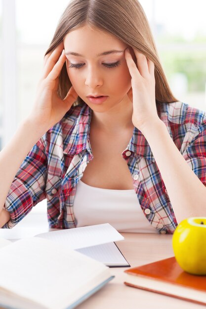 Sentirse cansado. Adolescente frustrada sosteniendo la cabeza entre las manos mientras está sentado en el escritorio con libros sobre ella