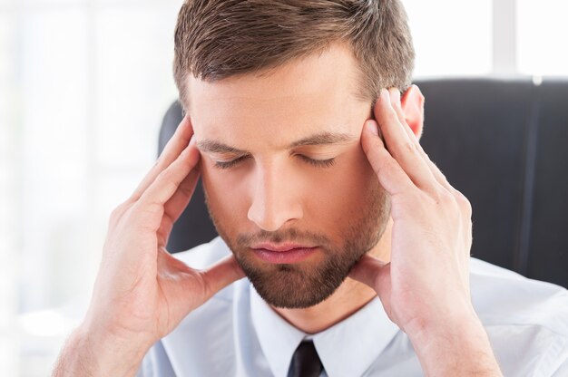 Sentirse cansado y deprimido. Hombre joven deprimido en camisa y corbata sosteniendo la cabeza entre las manos y manteniendo los ojos cerrados mientras está sentado en su lugar de trabajo