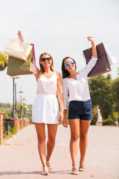 Sentirse feliz después del día de compras. Longitud total de dos hermosas mujeres jóvenes mostrando sus bolsas de la compra y sonriendo mientras caminan juntos por la calle